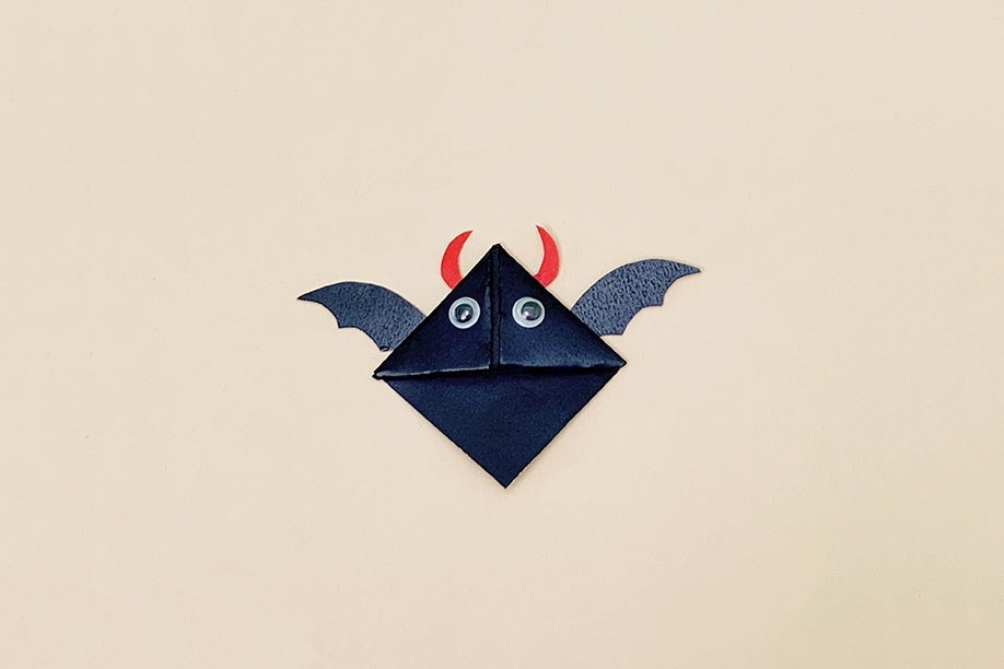 A completed origami bat bookmark is presented, showcasing a charmingly spooky design. The dark blue folded card forms the bat's body, while small black paper wings and red felt horns add to its character. Two white googly eyes are affixed to the front, giving the bat a friendly yet slightly mischievous expression. The bookmark is displayed centrally against a plain, off-white background, allowing its unique features to stand out clearly. The overall effect is playful and eye-catching, perfect for adding a touch of Halloween fun.