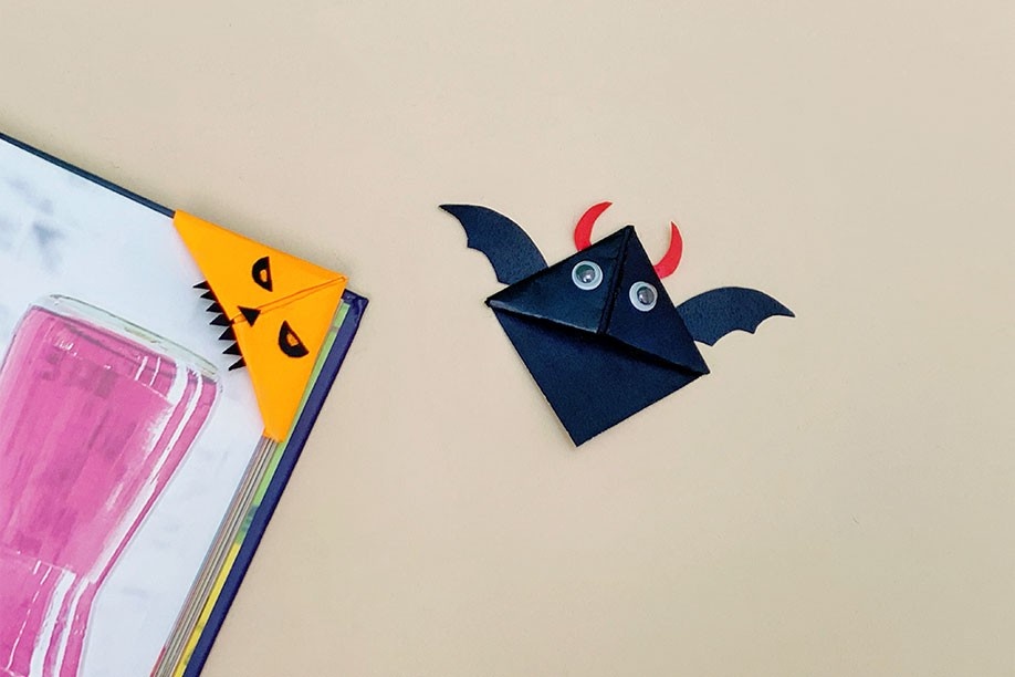 Two charming Halloween-themed corner bookmarks are displayed against a pale beige background. One, fashioned from orange card, depicts a playful monster face with jagged teeth and simple eyes. The other, a black bat, features small black wings and red horns, with white googly eyes adding a touch of whimsy. The orange bookmark is shown nestled in the corner of a book, showcasing its functionality. The overall impression is one of fun, simple crafts, perfect for adding a spooky touch to any book.