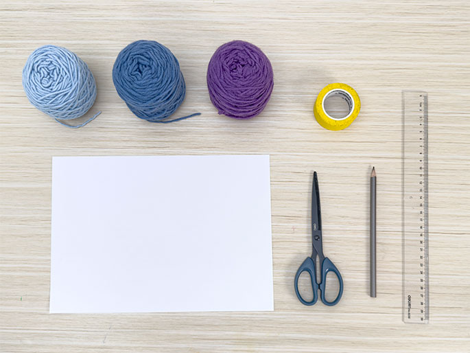 An assortment of craft supplies is arranged on a light woodgrain surface. Three balls of yarn in varying shades of blue and purple sit at the top. Below them are a blank white sheet of paper, a pair of scissors with dark handles, a pencil, a roll of yellow tape, and a clear plastic ruler. The image suggests preparation for a crafting project.