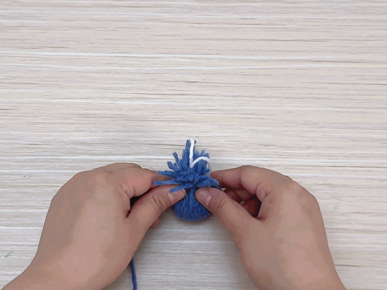 Two hands are working with a small, fluffy pom-pom made of blue yarn against a light woodgrain background. A piece of white yarn is visible near the top of the pom-pom, suggesting it's being used to tie and secure the shape. The hands are holding the pom-pom gently, likely in the process of tightening the white yarn or adding finishing touches.