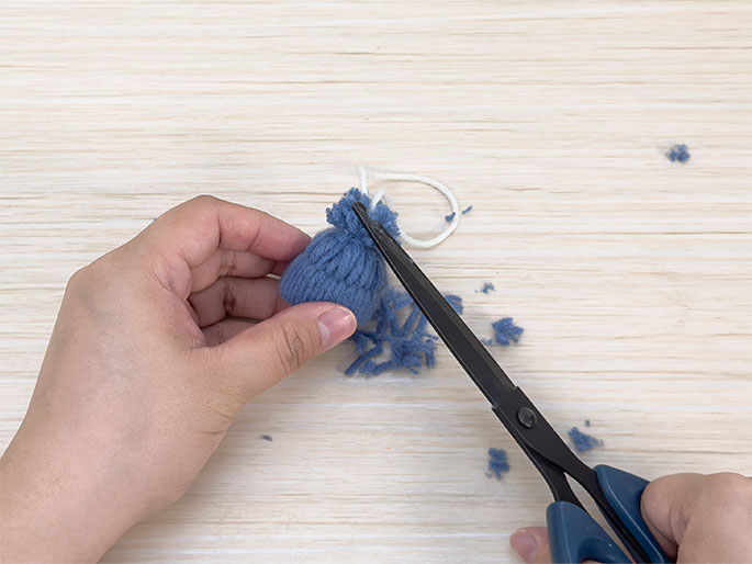 Against a light woodgrain background, one hand holds a small, blue yarn pom-pom tied with white yarn. Another hand uses a pair of blue-handled scissors to trim the uneven edges of the pom-pom. Small bits of trimmed blue yarn are scattered around the pom-pom on the surface. This image captures the final stages of making a neat, finished pom-pom.