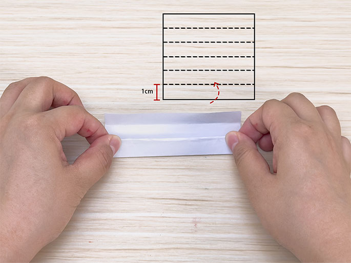 Two hands fold a strip of white paper in half lengthwise against a light woodgrain background. In the upper right corner, a diagram illustrates the folding process. The diagram shows a square divided into horizontal sections with dashed lines, and a red curved arrow indicates folding the bottom 1cm section upwards.