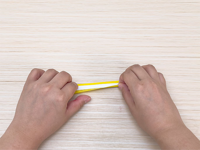 Two hands hold a thin strip of yellow tape with a white backing strip against a light woodgrain background. The hands are gently pulling the tape, likely to separate the backing from the adhesive side.