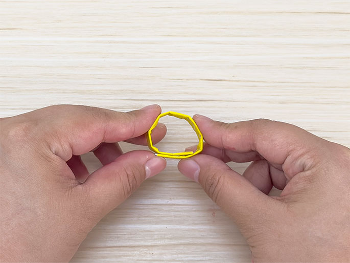 Two hands hold a small, folded piece of bright yellow material, possibly tape or paper, formed into a slightly irregular octagonal shape against a light woodgrain background. The object has a small opening in the center.