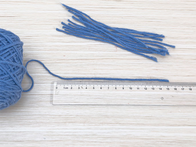 On a light woodgrain surface, a ball of blue yarn sits in the bottom left corner, with a strand extending to the right alongside a clear plastic ruler. The strand measures approximately 15cm. Above the ruler, a separate bundle of shorter, cut pieces of the same blue yarn is arranged. This suggests materials being prepared for a craft project, likely involving yarn tassels or fringe.