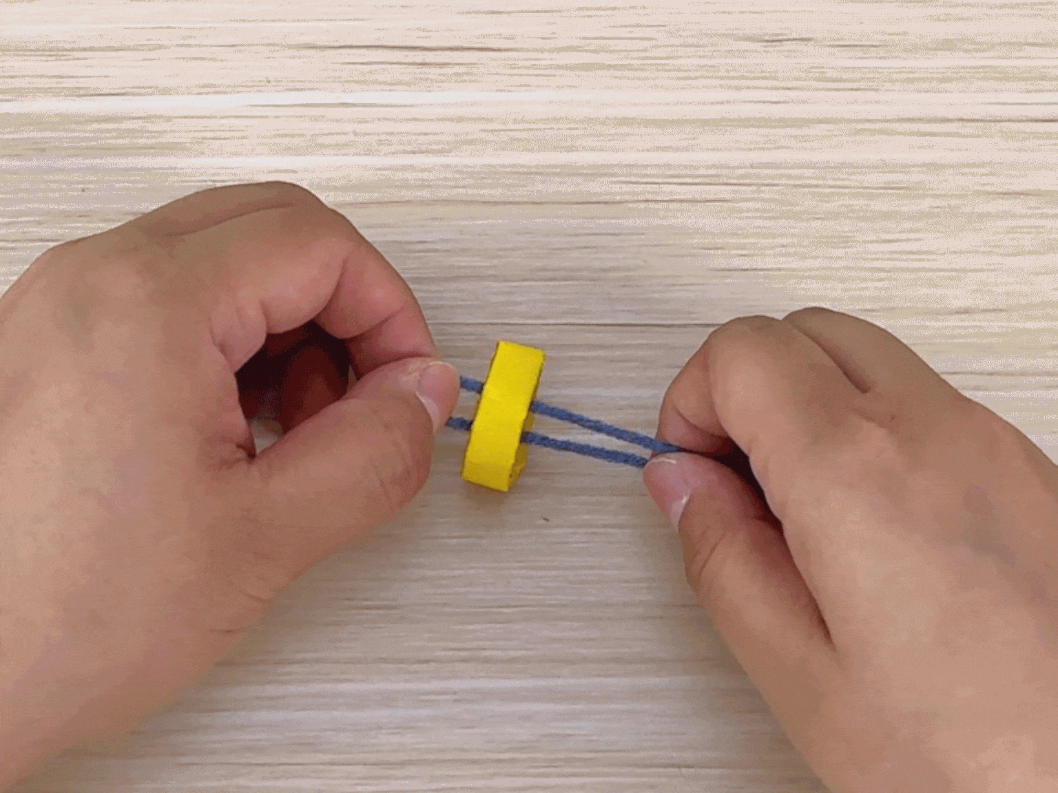 Against a light woodgrain background, two hands are shown manipulating a piece of blue yarn around a small, bright yellow ring or disc. The yarn is doubled over, and the hands appear to be in the process of tying or wrapping it around the yellow object. This suggests a crafting activity, possibly the initial steps in making a tassel, pom-pom, or other yarn-based decoration.