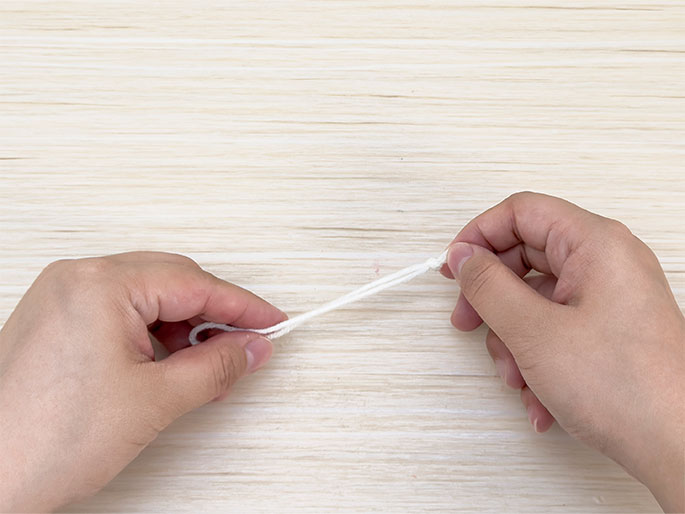 Two hands hold a length of white yarn or string against a light woodgrain background. A simple overhand knot has been tied near the right hand, and a small loop is formed near the left hand. The hands appear to be gently pulling the yarn taut. This image likely depicts a stage in a crafting or knot-tying process.
