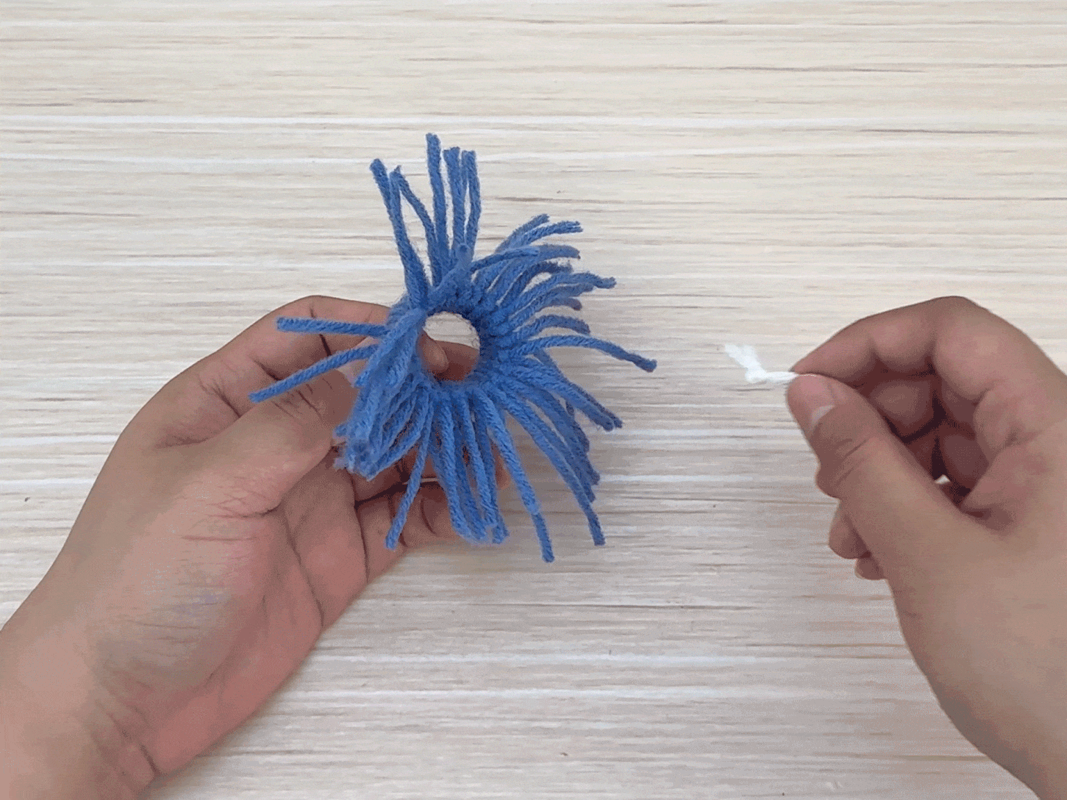 On a light woodgrain background, a hand holds a small, ring-shaped object covered in short strands of blue yarn, resembling a pom-pom or tassel. Another hand, on the right side of the image, holds a short piece of white yarn or string. This appears to be a craft project in progress, possibly involving the creation of a yarn-based decoration.
