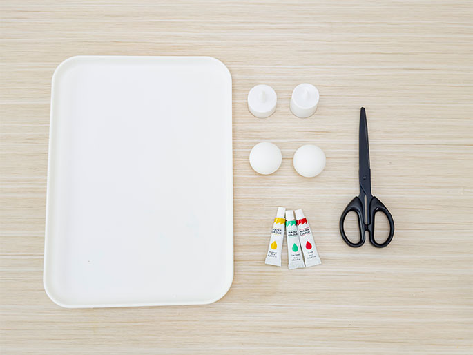 An overhead flatlay image shows various craft supplies arranged on a light wood grain surface. A rectangular white tray occupies the top left corner. To its right, two small white LED tea lights are positioned above two white polystyrene balls. Below the balls are three small tubes of watercolor paint in yellow, green, and red. A pair of black scissors lies to the right of the paints. The arrangement suggests preparation for a craft project involving painting the polystyrene balls.
