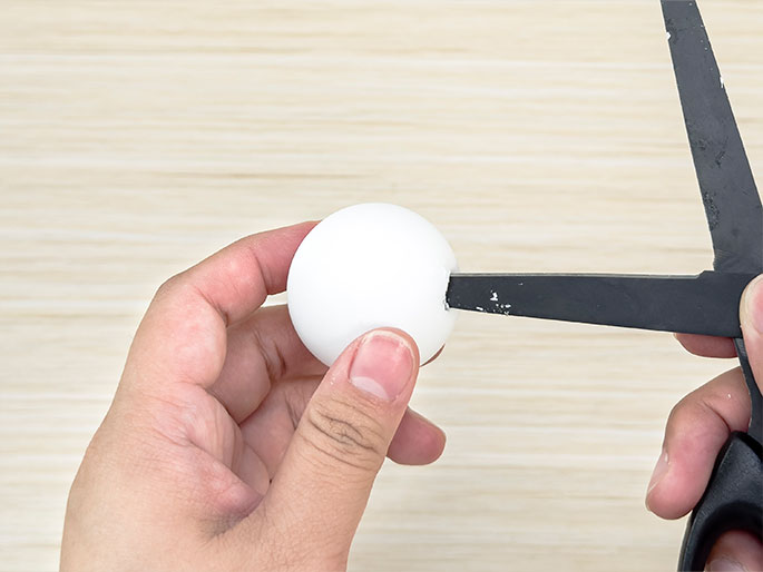Two hands are shown manipulating a white polystyrene ball and a pair of scissors against a light wood-grain background. One hand holds the ball securely while the other hand uses the scissors to make a small cut or notch in the ball's surface. The focus is on the process of modifying the ball with the scissors.