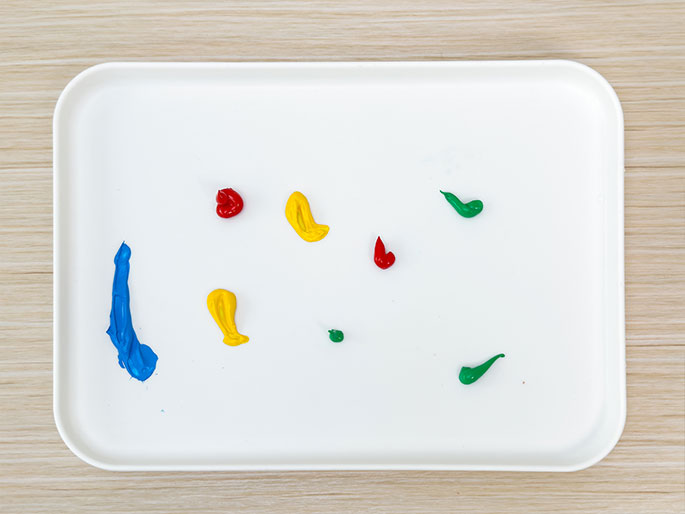 A white rectangular tray sits on a light wood-grained surface. Several small dabs of paint in red, yellow, blue, and green have been squeezed onto the tray's surface. The dabs vary slightly in size and shape. The image suggests an overhead view of a simple painter's palette ready for use.