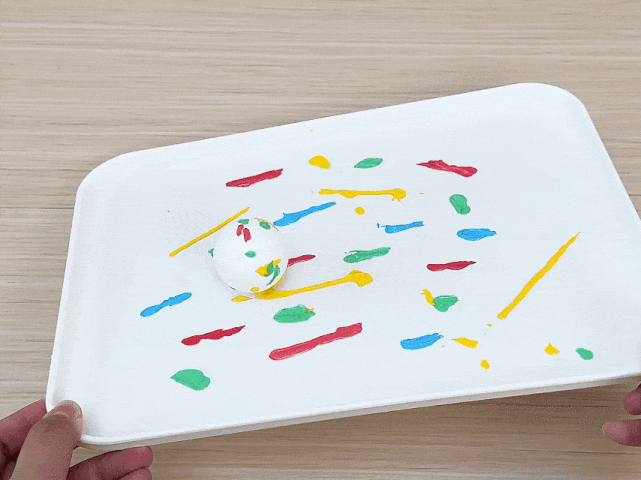 Two hands hold a white rectangular tray above a light wood-grained surface. On the tray, a white ball, partially decorated with streaks and dabs of red, yellow, blue, and green paint, rests amidst more paint streaks and splatters in the same colors. The scene appears to depict a work in progress, likely a craft project involving decorating the ball.
