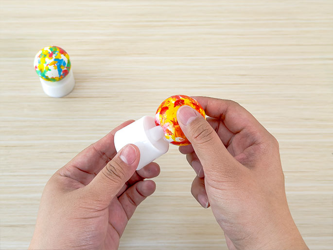 Against a light wood-grain background, two hands are shown assembling a small decorative light. One hand holds a white cylindrical base, possibly an LED tea light, while the other hand positions a small sphere decorated with orange and yellow paint onto the base. In the background, another similarly decorated sphere already sits atop a base. The image captures the process of creating these small, colorful lights.