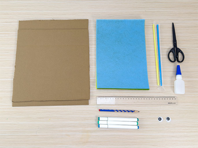 An overhead flatlay shows the materials needed for a craft project arranged on a light wood grain surface. The materials include two pieces of brown cardboard, sheets of light blue and bright green felt or foam, yellow, light green, and blue straws, a pair of scissors, a bottle of white glue, a clear plastic ruler, a pencil, three white marker pens, and a pair of googly eyes. These items are neatly laid out, ready for use.