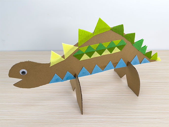 A completed cardboard dinosaur craft stands on a light wooden table. The dinosaur, made from brown cardboard, is in profile facing left and has two legs for support. It's decorated with colorful felt triangles: light yellow and various shades of green triangles form a spiky ridge along its back, and light blue triangles line its underbelly. A googly eye is attached to the dinosaur's head. The craft appears to be a children's project.