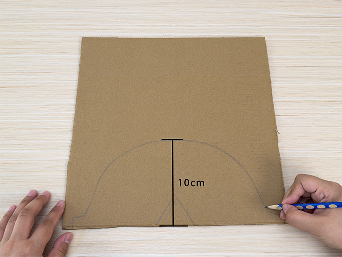 On a light wooden surface, a piece of brown cardboard has a partially drawn shape sketched onto it in pencil. The shape resembles a rounded arch with a small triangular point at the bottom center. A hand holding a blue pencil is in the process of drawing the right side of the arch. A vertical line with 10cm written next to it indicates the height of the arched shape. Two hands are visible, one holding the cardboard steady while the other draws. This image appears to show the first steps in creating a cardboard cutout.