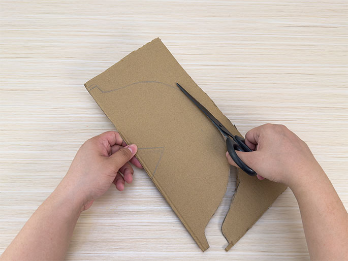 Two hands are shown cutting a piece of brown cardboard on a light wood grain surface. One hand holds the cardboard while the other uses scissors to cut along a pre-drawn curved line. Part of the curved shape has already been cut away. The remaining portion of the cardboard still has visible pencil markings.