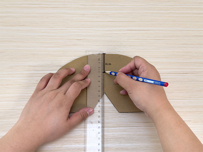 On a light wood-grain surface, two hands are shown working with a piece of brown cardboard shaped like a semi-circle with a triangular notch cut out at the bottom. A clear plastic ruler is placed vertically along the center of the notch, and one hand holds a blue pencil to mark a point on the cardboard at the 4cm mark on the ruler. The other hand holds the cardboard and ruler steady. A small vertical line next to the 4cm mark indicates the measurement being made. This image appears to depict a step in a crafting project involving precise measurements.
