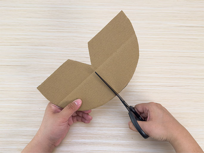 Two hands are shown cutting a piece of folded brown cardboard with scissors on a light wood-grain surface. The cardboard is folded in half, creating a curved shape on one side and an angled edge on the other. The scissors are cutting along the folded edge. One hand holds the cardboard while the other manipulates the scissors.