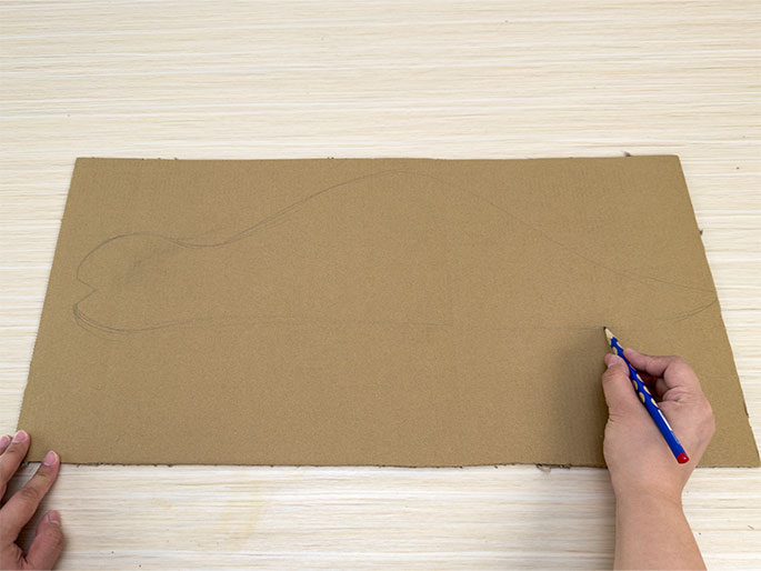 On a light wooden surface, a rectangular piece of brown cardboard lies flat. A hand holding a blue pencil is drawing a curved, fish-like shape on the cardboard. Another hand is partially visible, holding the bottom left corner of the cardboard. The pencil sketch is still in progress, with only the outline of the fish being drawn.
