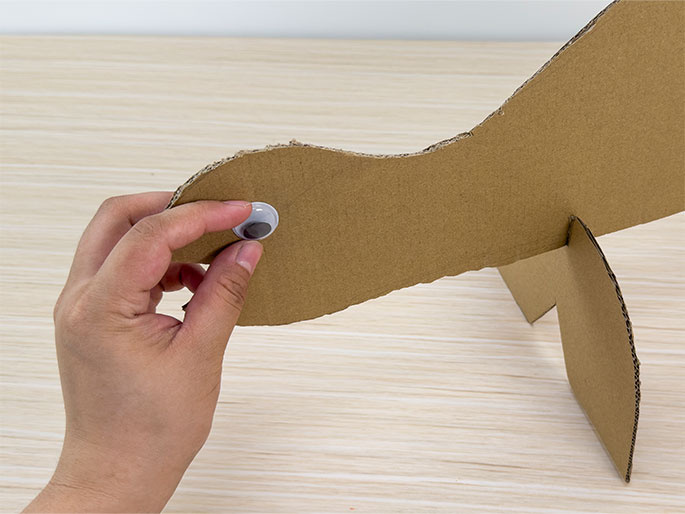 A hand is shown attaching a googly eye to a cardboard dinosaur cutout on a light wood-grain table. The dinosaur, made of brown cardboard, is positioned with its profile facing left and is supported by two cardboard legs. The hand is holding the plastic googly eye against the dinosaur's head. The focus is on the act of adding the eye to the cutout.