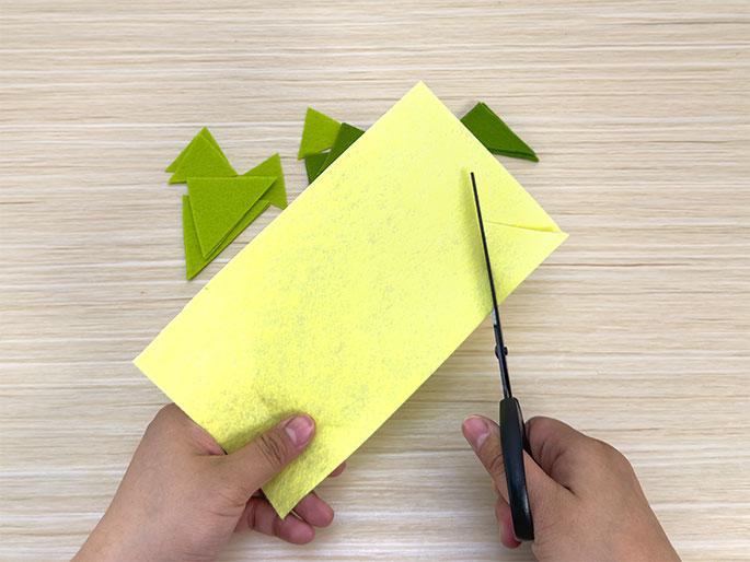 Two hands are shown working with pieces of felt on a light wood-grain surface. One hand holds a rectangular piece of light yellow felt, while the other hand uses black scissors to cut a small triangle from the corner of the felt. Several small, pre-cut green triangles are visible near the top left of the image.