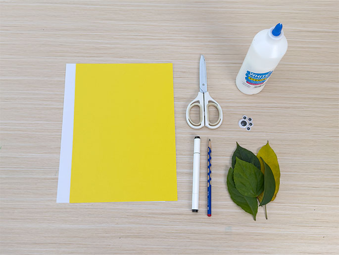 An assortment of craft supplies lies on a light wooden table, arranged for a flat lay photograph. A large sheet of bright yellow construction paper is positioned on the left. To the right are a pair of small, white-handled scissors, a bottle of white glue, a set of small, stick-on googly eyes, a white marker, a pencil, and a cluster of green and yellow-green leaves. These materials suggest preparation for a nature-themed craft project.