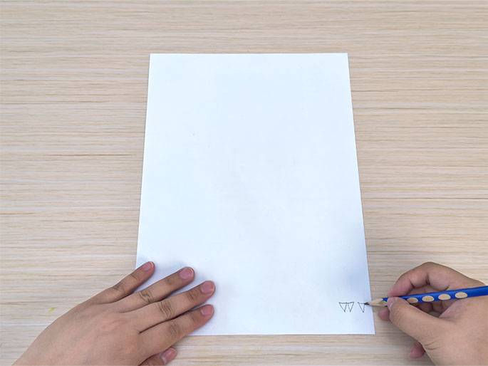 A hand draws small, connected triangles along the bottom edge of a blank white sheet of paper. Another hand holds the paper steady on a light wood grain table. The hand drawing uses a short pencil with blue and white dots. This image likely depicts the beginning stages of a drawing or craft project.