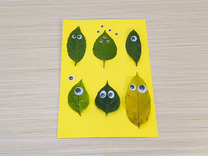 Six leaves are arranged on a bright yellow piece of paper, which sits on a light wood grain surface. Each leaf has googly eyes attached, giving them whimsical faces. The leaves are in two rows of three. The top row has three green leaves, each with two eyes. The bottom row has a lighter green leaf with two eyes, a darker green leaf with two eyes, and a yellow leaf with two eyes. A few extra googly eyes are scattered on the yellow paper near the top. This image likely depicts a children's craft project.
