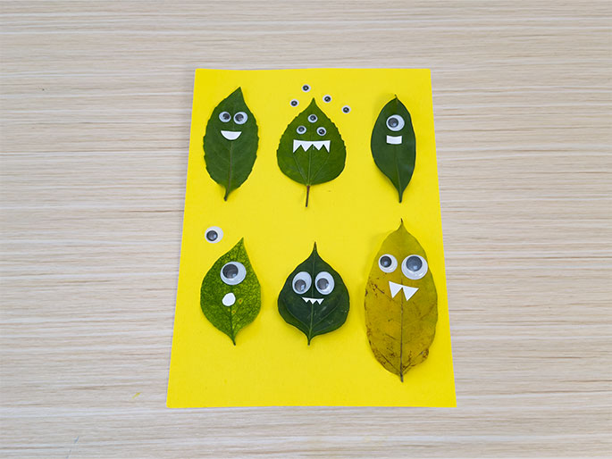 Six leaves are arranged on bright yellow paper, set on a light wood grain surface. Each leaf has been transformed into a whimsical monster face using googly eyes and drawn mouths. The leaves are arranged in two rows of three. The top row features three green leaves. The first has two eyes and a smile. The second has three eyes and jagged teeth. The third has two eyes and a small, straight mouth. The bottom row includes a lighter green leaf with two eyes and a surprised o shaped mouth, a darker green leaf with two eyes and jagged teeth, and a yellow leaf with two eyes and jagged teeth. A few extra googly eyes are scattered on the yellow paper near the top. This image likely depicts a finished children craft project.