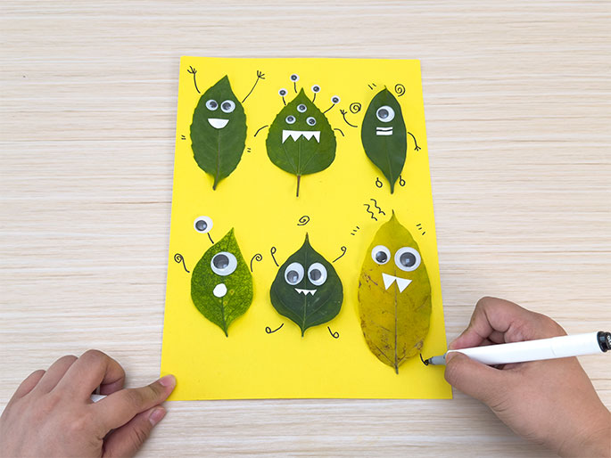 Leaf monsters with googly eyes and drawn features on yellow paper. A hand adds details with a marker.