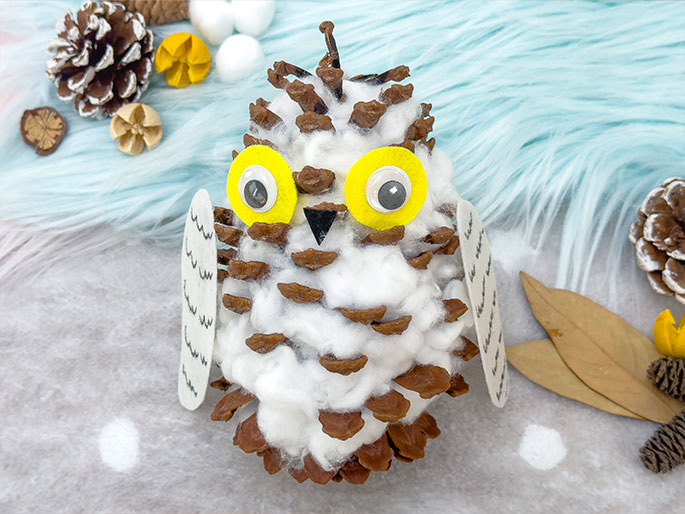 A charming handmade owl craft is displayed on a textured, light blue and white background. The owl's body is a pine cone, covered with fluffy white cotton. It has large, yellow felt circles for eyes, each with a smaller black googly eye glued to the center. Two white, wing-shaped pieces of material, decorated with wavy lines resembling feathers, are attached to the sides of the pine cone. Small craft supplies like other pine cones and dried leaves are scattered around the owl.