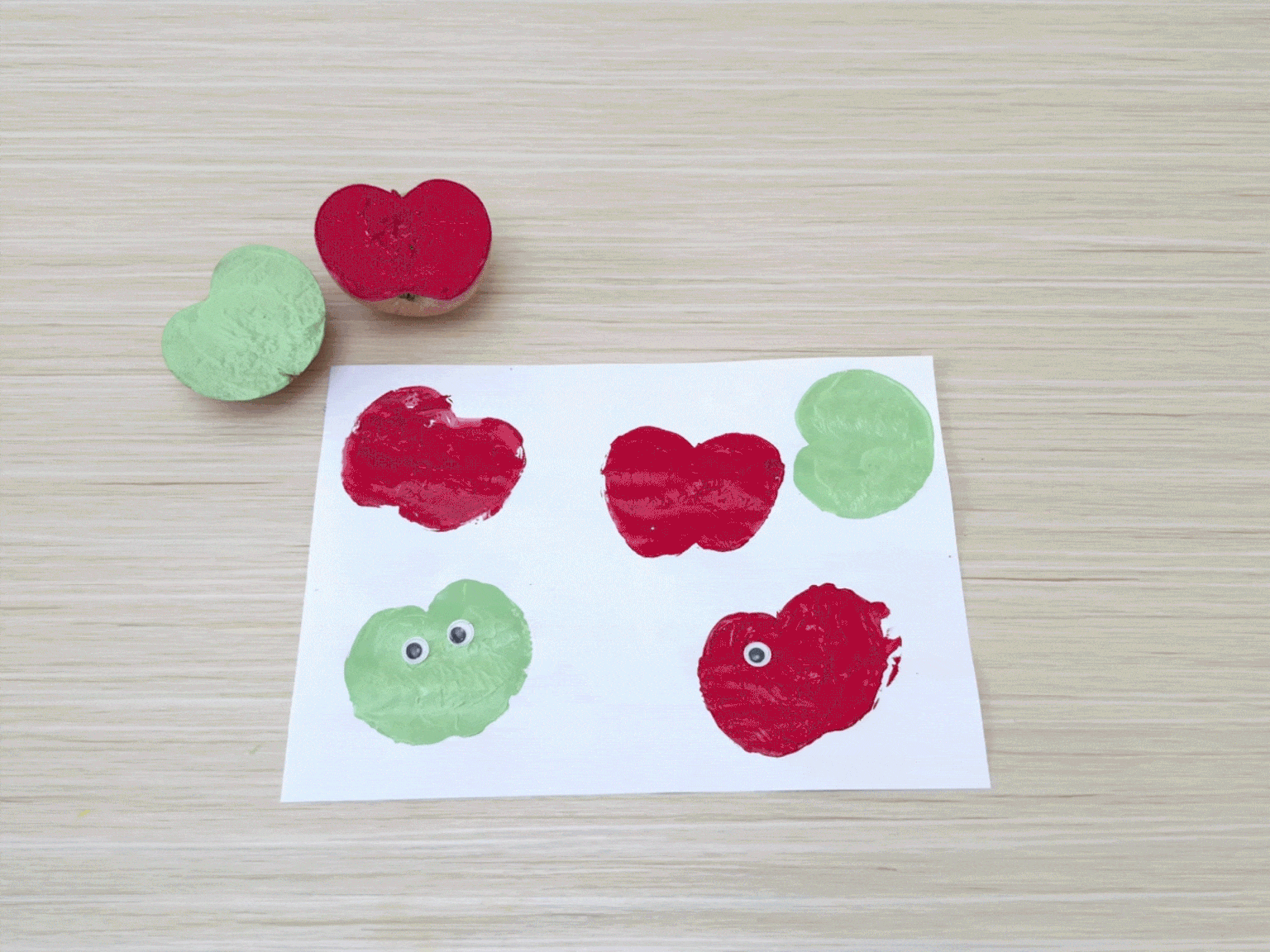 A piece of white paper on a light wood-grain table displays four apple prints made using halved apples as stamps. Two prints are red, and two are light green. Each print has two small googly eyes attached, giving them a cartoonish appearance. Simple, hand-drawn arms and legs are added to the bottom two apple prints, making them look like they are dancing. Two apple halves, one red and one green, sit at the top left corner of the paper, showing the tools used to create the prints. The video showcases a simple and fun apple stamping art activity.