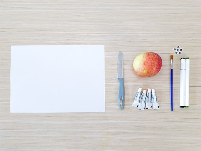 Art supplies are arranged on a light woodgrain table, suggesting preparation for an art project. A blank sheet of white paper sits on the left. A small paring knife with a gray handle lies to the right of the paper, followed by a red and yellow apple. Next to the apple are several small tubes of paint or glue, a paintbrush with a blue handle, and two markers or pens. This flat lay composition indicates the materials gathered for a creative activity, likely involving the apple as a subject.