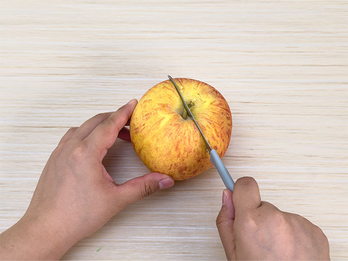 Two hands are shown on a light woodgrain surface, in the process of cutting an apple. One hand holds the apple steady while the other hand uses a small paring knife with a gray handle to slice into it. The apple is red and yellow and the knife is positioned near the top center of the fruit. The focus is on the action of cutting the apple.