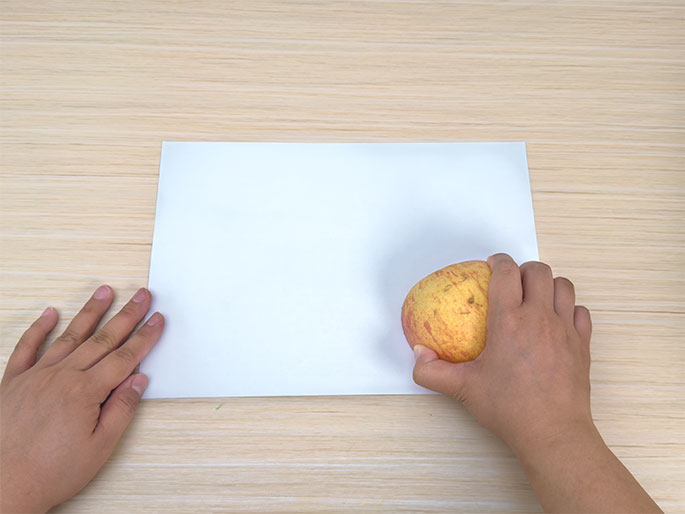 Two hands are positioned above a blank sheet of white paper on a light woodgrain table. One hand rests lightly on the edge of the paper, while the other hand holds a red and yellow apple. The apple is positioned near the paper, as if about to be used for printing or stamping. The scene is set for an art activity, likely apple printing.