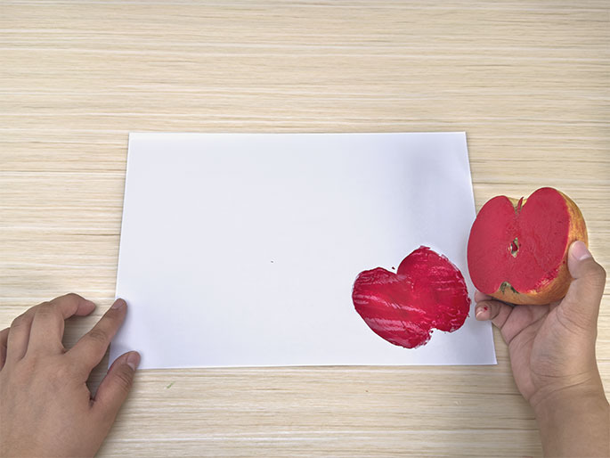 On a light woodgrain table, a piece of white paper displays a fresh, bright red apple print. A hand holds a cut half of an apple, also coated in red paint, just above the paper, suggesting the apple was used to create the print. Another hand is visible, lightly touching the paper. The scene clearly depicts apple printing in progress.