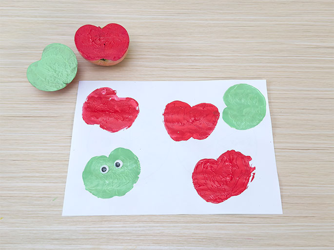 A white sheet of paper on a light woodgrain table displays several apple prints made with red and green paint. Two cut apple halves, one red and one green, sit above the paper, indicating the tools used for the printing. One of the green apple prints has googly eyes attached, creating a simple face. The arrangement showcases a finished apple printing craft project.