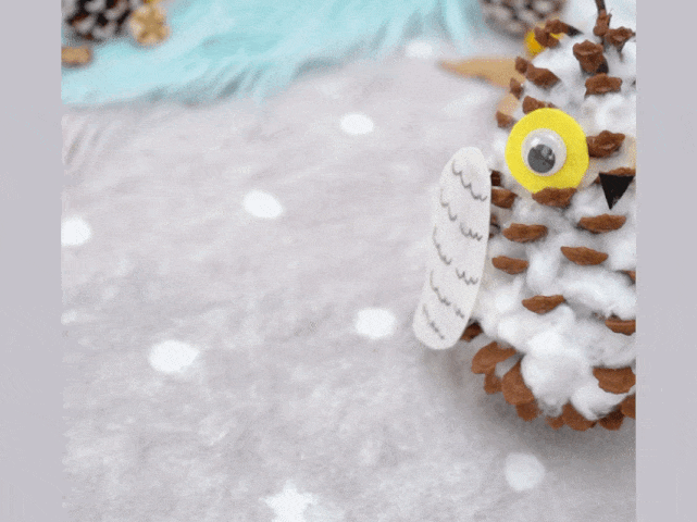 A hand-crafted owl, made from a pine cone, is displayed on a textured white surface. The pine cone forms the owl's body, covered with fluffy white cotton balls, likely representing feathers. Large, yellow googly eyes and a small, triangular beak give the owl a friendly appearance. Small, white paper wings are attached to the sides. The background includes other natural elements, possibly more pine cones and some fabric, suggesting an autumnal or nature-themed craft project.