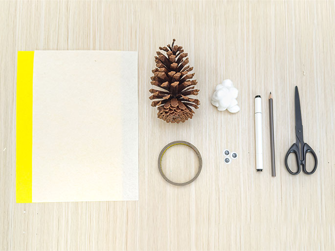 An overhead flatlay image displays various craft supplies arranged on a light wood grain surface. A sheet of off-white paper with a vertical bright yellow stripe along one edge sits on the left. To its right, a brown pine cone lies next to a small clump of white crafting clay or cotton. A roll of clear tape is positioned below the pine cone. To the right, two small googly eyes, a white marker, a brown pencil, and a pair of black scissors are arranged in a line. The items suggest preparation for a nature-themed craft project.