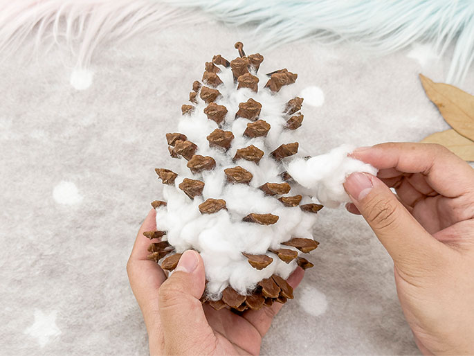 Two hands are working on a craft project against a soft, textured background of white and pastel colors. One hand holds a pine cone, while the other hand applies fluffy white cotton wool between the cone's scales. The process appears to be creating a snowy or winter-themed decoration using the pine cone as a base.