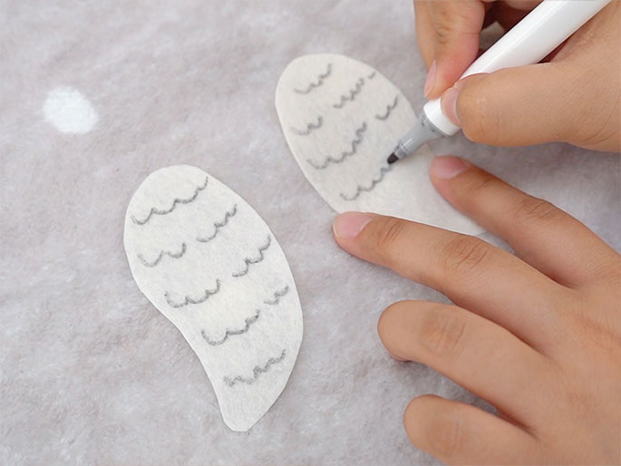 On a textured, off-white surface, two wing-shaped pieces of thin white material lie slightly apart. A hand holding a white marker is drawing wavy lines onto one of the wing shapes, creating the appearance of feathers. The other wing shape has similar lines already drawn on it. The scene suggests a craft project involving creating decorative wings.