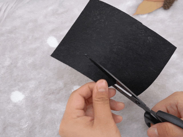 A pair of hands uses black scissors to cut a piece of black paper or fabric. The scissors are making a cut from one edge of the material, which is positioned against a textured off-white background with faint lighter circles. The focus is on the action of cutting the dark material.