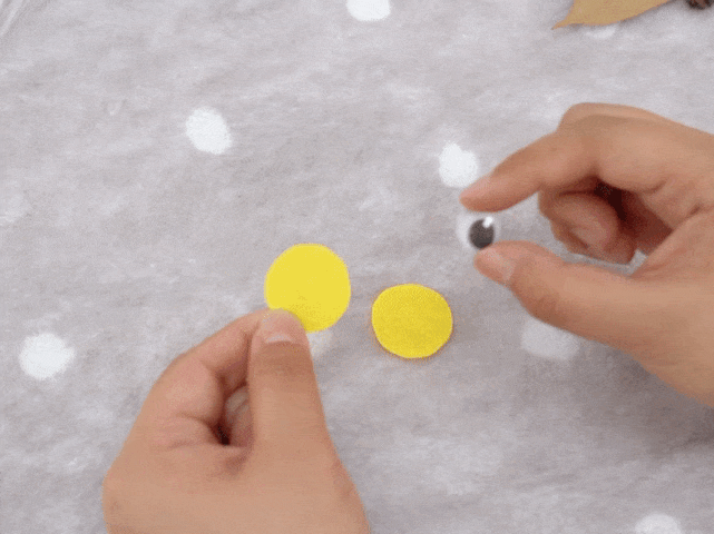 Two hands hold small craft supplies against a textured, off-white background with faint lighter circles. One hand holds two small, bright yellow circles of felt or fabric. The other hand holds a googly eye. The arrangement of the materials suggests the next step in a craft project, likely attaching the eye to the yellow circles.
