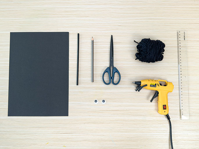 Arranged on a light wood-grain surface are the materials for a craft project. From left to right, there's a sheet of black construction paper, a black straw or stick, a pencil, a pair of blue-handled scissors, two googly eyes, a ball of black yarn or string, a yellow hot glue gun, and a clear plastic ruler. These items appear to be laid out in preparation for assembly.