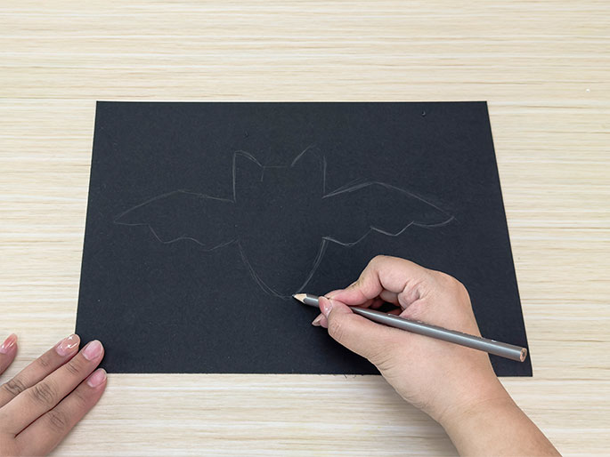 On a piece of black construction paper, a hand draws the outline of a bat with a pencil. The bat is positioned centrally with its wings spread, and the pencil is actively tracing the lower portion of the bat's body. The background is a light wood-grain surface, and a second hand is partially visible at the lower left, holding the paper in place.