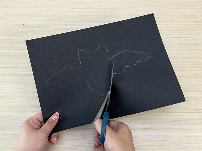 A pair of hands is cutting out a bat shape from black construction paper. The bat outline, drawn in pencil, is visible on the paper, and blue-handled scissors are in the process of cutting along the edge of one wing. The background is a light wood-grain surface.
