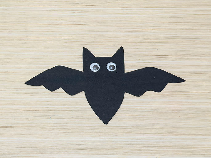 A cut-out of a black construction paper bat lies on a light wood-grain surface. The bat has outstretched wings, pointed ears, and two large googly eyes, giving it a simple, childlike appearance.