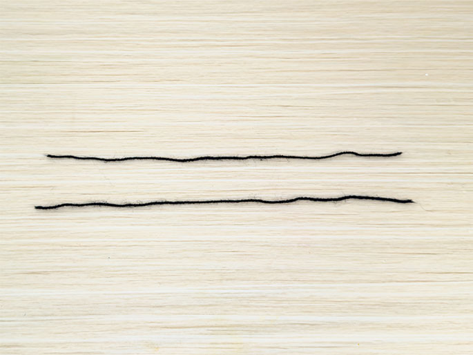 Two lengths of black yarn or string are laid parallel to each other on a light wood-grain surface. They are positioned horizontally across the center of the image, with a small gap between them.