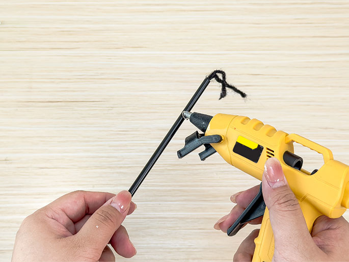 Two hands are shown working on a craft project against a light wood-grain background. One hand holds a black stick or straw, while the other hand operates a yellow hot glue gun. A small amount of black yarn is attached to the top of the stick, indicating that the glue gun is being used to adhere the yarn to the stick.